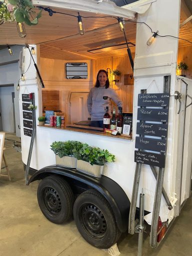 Eine mobile Bar im Anhänger mit einer Person, die Getränke serviert. Dekorative Pflanzen sind sichtbar.